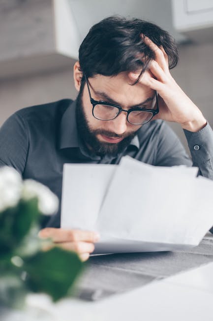 A worried man with glasses reviews financial papers, highlighting stress and financial concern.